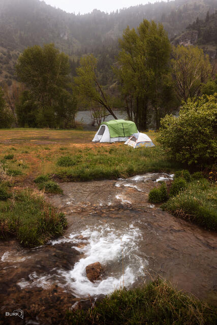 Two tents beside a flowing creek at Porcupine Reservoir in northern Utah, surrounded by green trees, mountain slopes, and calm water beyond.