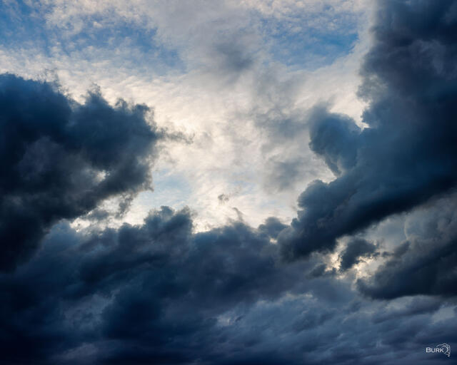Dramatic dark storm clouds surround a bright opening of blue sky and glowing light, creating a powerful and atmospheric cloudscape.