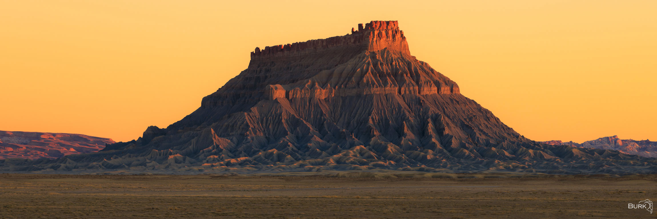 Factory Butte Alpenglow This panoramic photograph captures the striking presence of Factory Butte at sunrise, as a warm alpenglow gently illuminates...